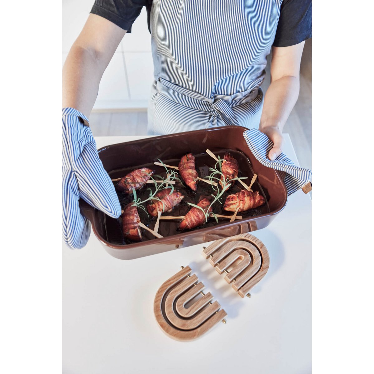 Person holding a baking dish with wrapped meat and rosemary, wearing oven mitts and an a blue denim apron.