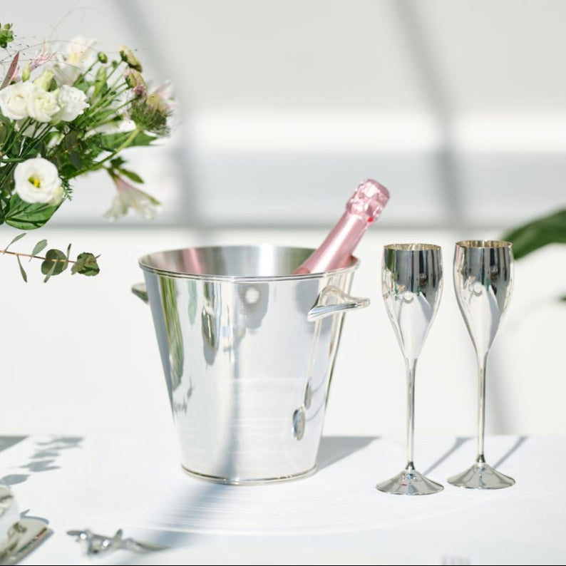 Silver champagne bucket with pink bottle and two flutes on a white surface with flowers in the background