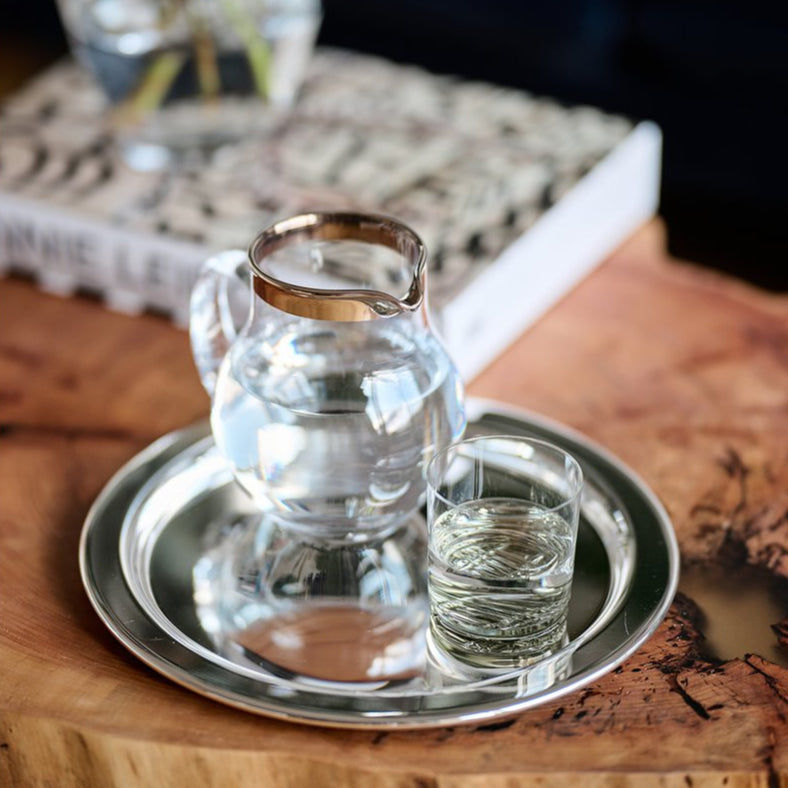 Clear glass pitcher and cup on a silver tray with a wooden surface and blurred background