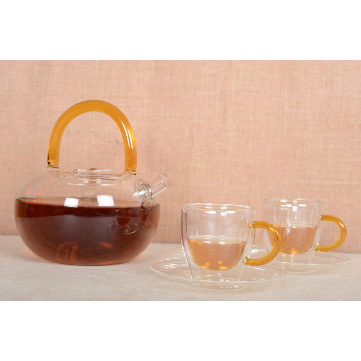 Glass teapot with amber handle and two matching glass cups on a beige background