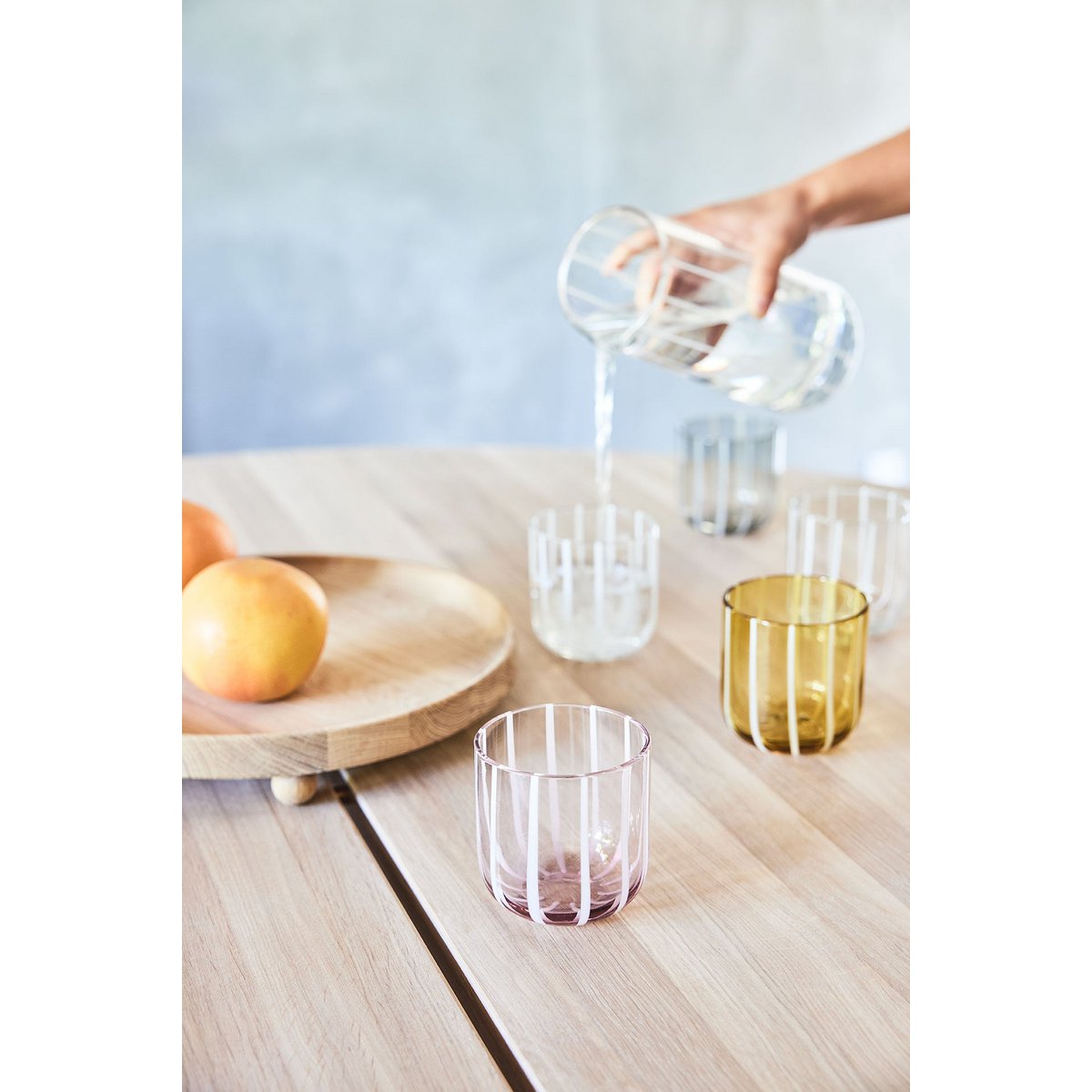 Clear glass pitcher being filled with water on a wooden table with colorful glasses.