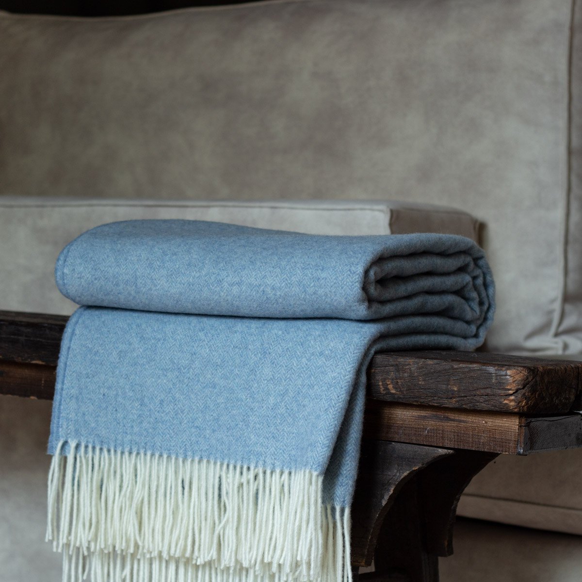 Folded blue blanket with white fringe on a wooden bench against a neutral background