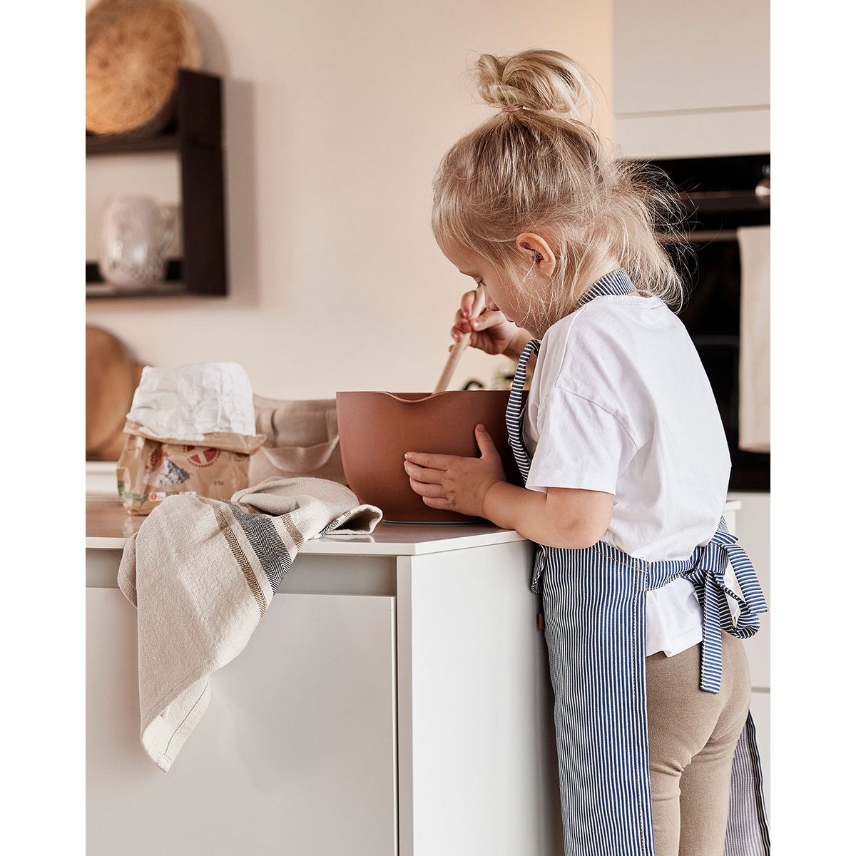 Child in a kitchen wearing an apron, standing at a counter with a brown bowl.