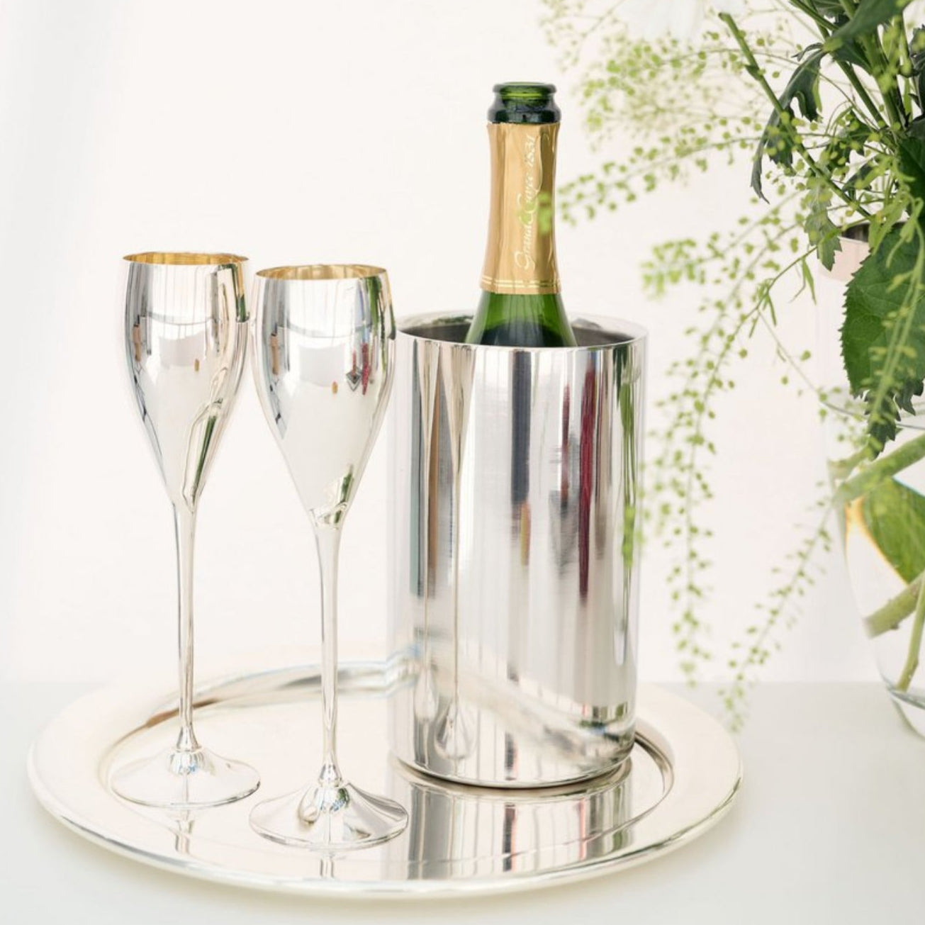 Silver champagne bucket with two flutes and a bottle of champagne on a white surface with flowers in the background.