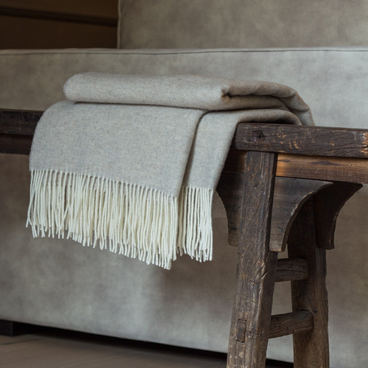 Gray blanket with white fringe draped over a wooden bench against a neutral background