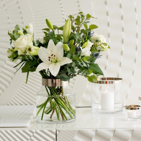 Bouquet of white flowers in a clear vase with three small candles on a white surface.
