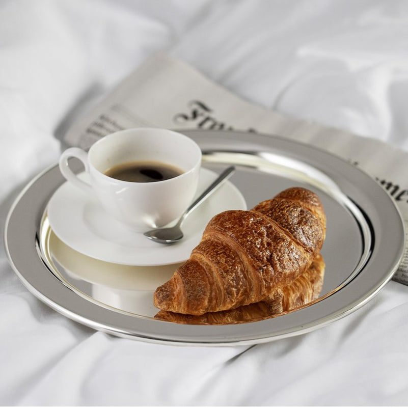Cup of coffee with a croissant on a silver tray on a white surface with a newspaper.