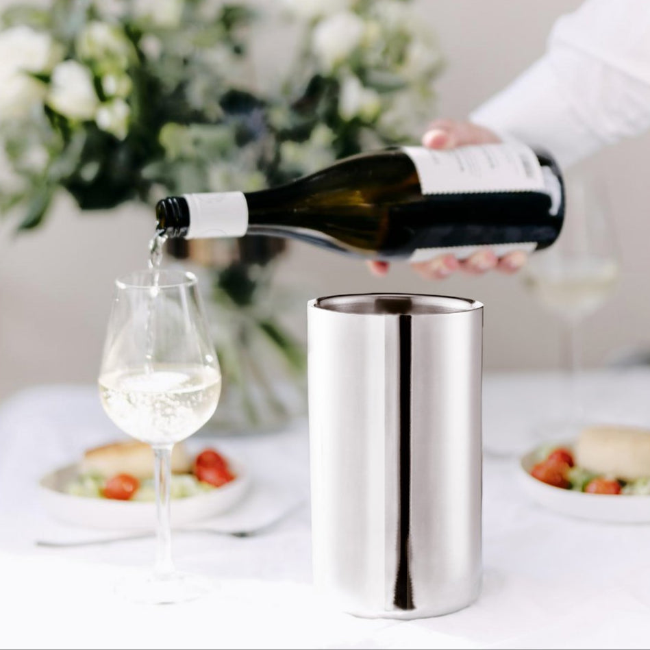 Person pouring white wine into a glass with a stainless steel cooler on a table.