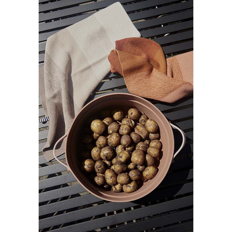 Brown ceramic pot filled with potatoes on a dark wooden surface with a beige towel and brown cloth.