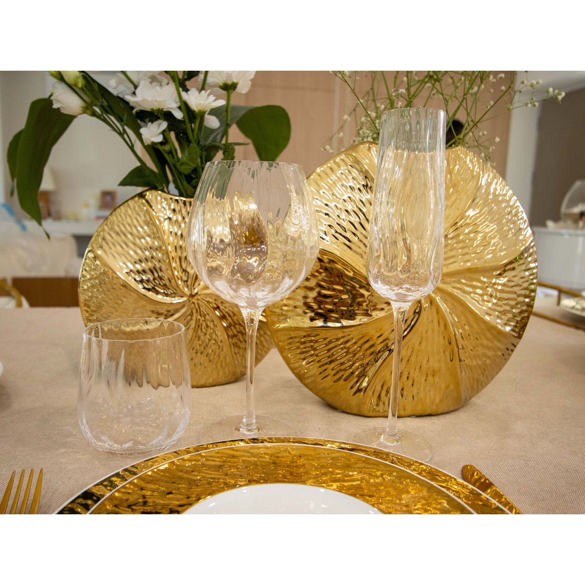 Dining table setting with gold plates and clear glasses on a neutral background