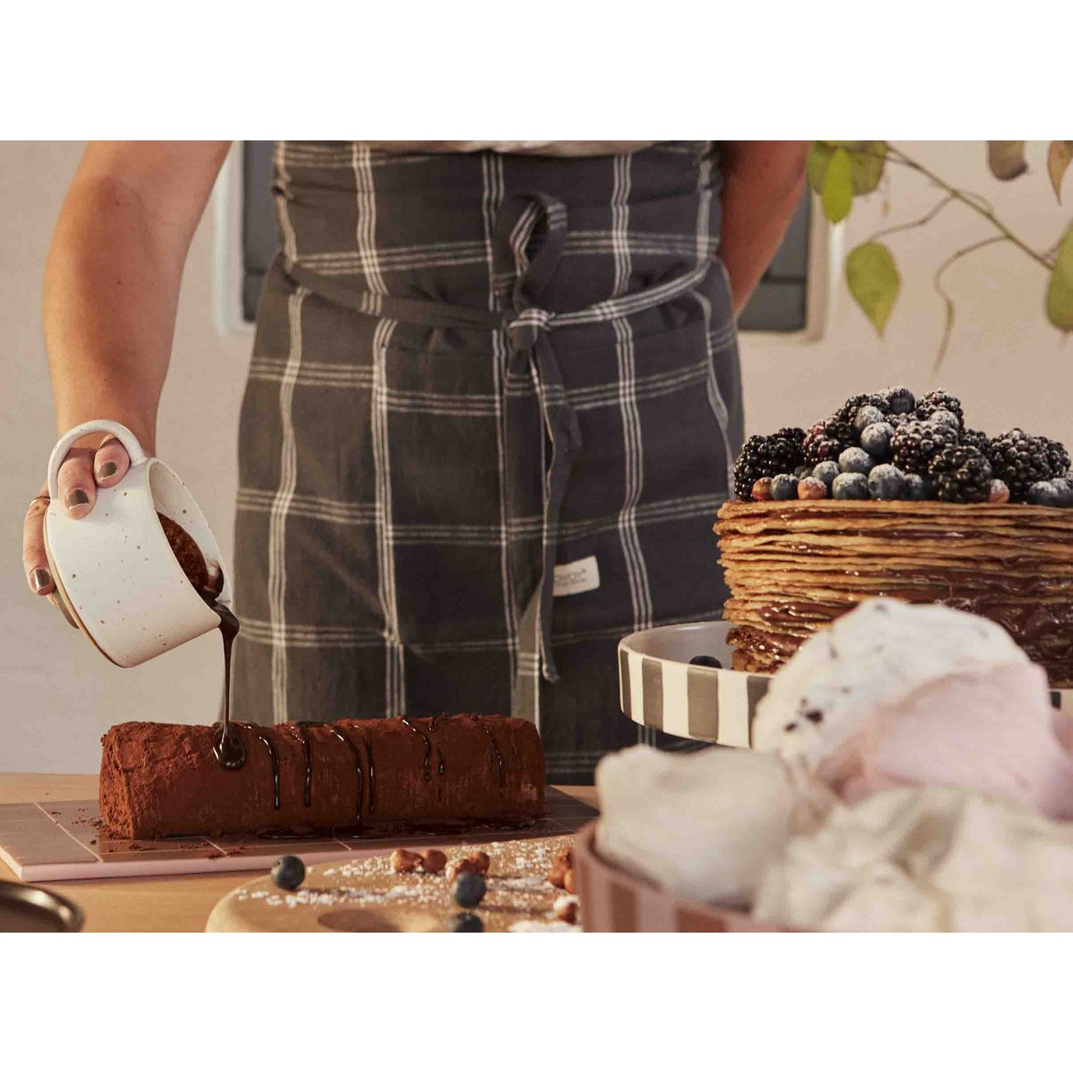 Person pouring chocolate onto a cake with a plaid apron, surrounded by berries and other desserts.