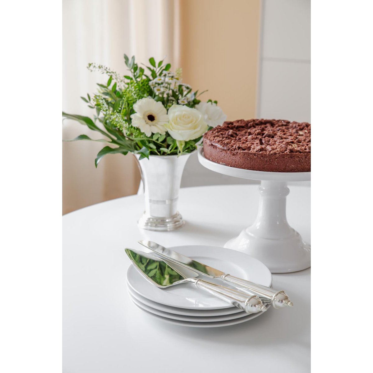 Table setting with white plates, silver knife, cake, and floral arrangement on a white surface.